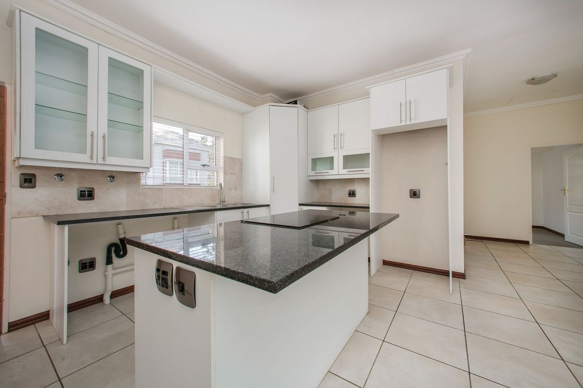 A bright, empty kitchen featuring white cabinetry, granite countertops, a central island with a built-in hob, and neutral ceramic floor tiles.
