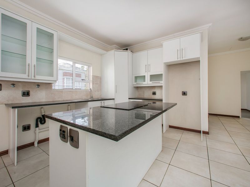 A bright, empty kitchen featuring white cabinetry, granite countertops, a central island with a built-in hob, and neutral ceramic floor tiles.
