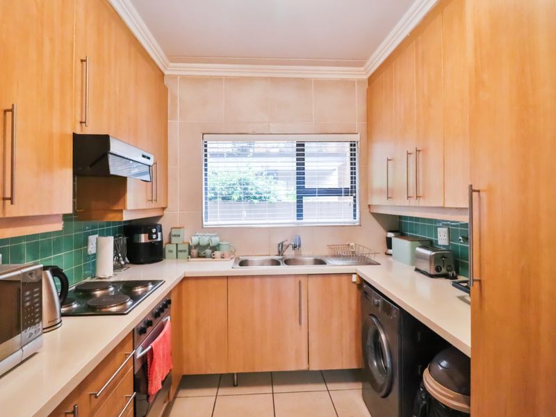 A functional galley-style kitchen featuring light wood cabinetry, tiled backsplash, integrated appliances, and a double sink with natural light from the window.