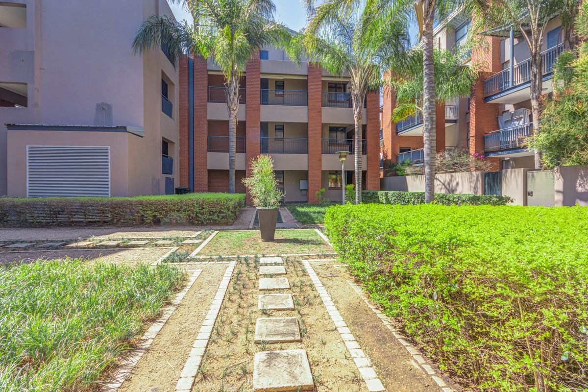 A well-maintained communal garden area featuring paved pathways, manicured hedges, and mature palm trees set against the brick facade of the apartment complex.