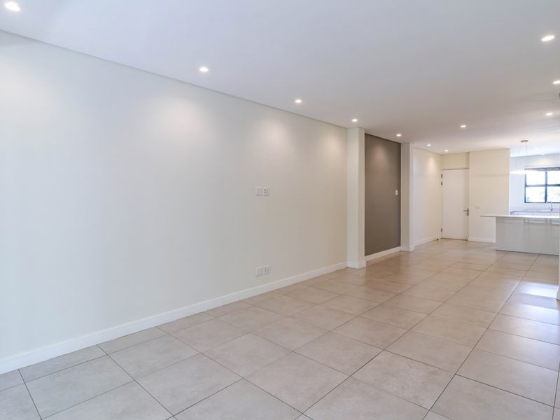 A bright, open-plan living and kitchen area featuring neutral tiled flooring, recessed downlighting, and a clean, modern aesthetic.