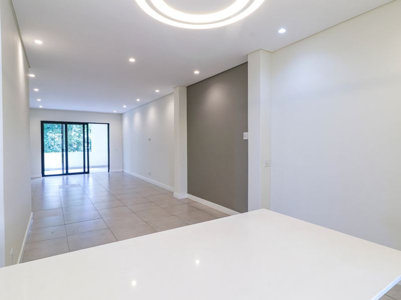 A bright, modern open-plan living area featuring neutral tiled floors, recessed lighting, an accent wall, and sliding glass doors leading to an outdoor space.