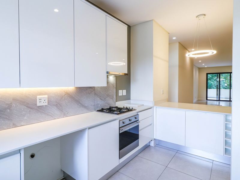 Modern kitchen featuring white high-gloss cabinetry, stone countertops, integrated gas hob, oven, and under-cabinet lighting, flowing into an open-plan living area.
