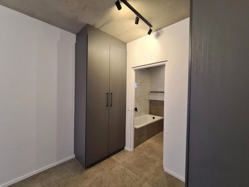 Modern bedroom featuring built-in cupboards, exposed concrete ceiling with track lighting, and direct access to an en-suite bathroom with tiled finishes.