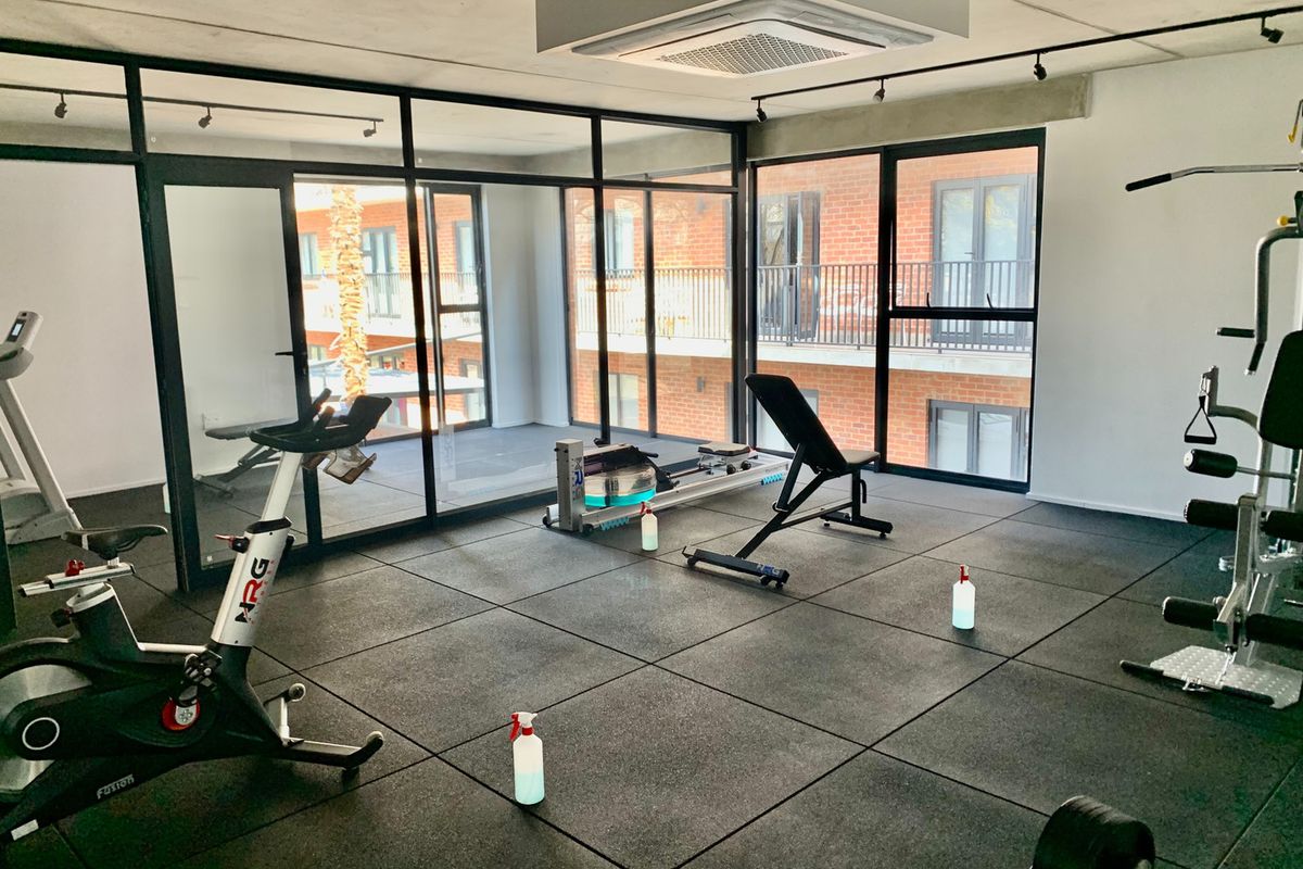 A modern, well-maintained gym facility featuring rubber flooring, track lighting, and floor-to-ceiling glass partitions overlooking the apartment complex.