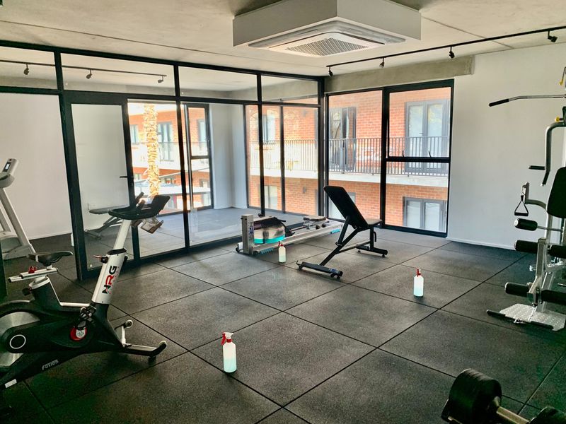 A modern, well-maintained gym facility featuring rubber flooring, track lighting, and floor-to-ceiling glass partitions overlooking the apartment complex.