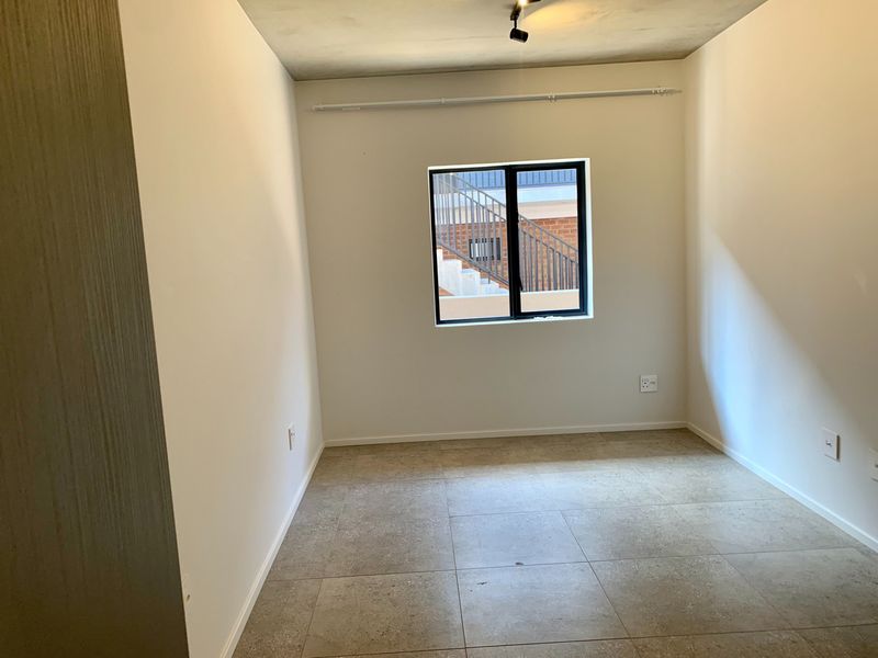A clean, empty bedroom featuring neutral tiled flooring, an exposed concrete ceiling with track lighting, and a modern black-framed window.