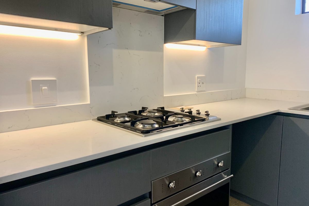 Modern kitchen featuring dark cabinetry, stone countertops, a gas hob, built-in oven, and integrated extractor fan with under-cabinet lighting.