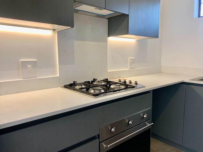Modern kitchen featuring dark cabinetry, stone countertops, a gas hob, built-in oven, and integrated extractor fan with under-cabinet lighting.