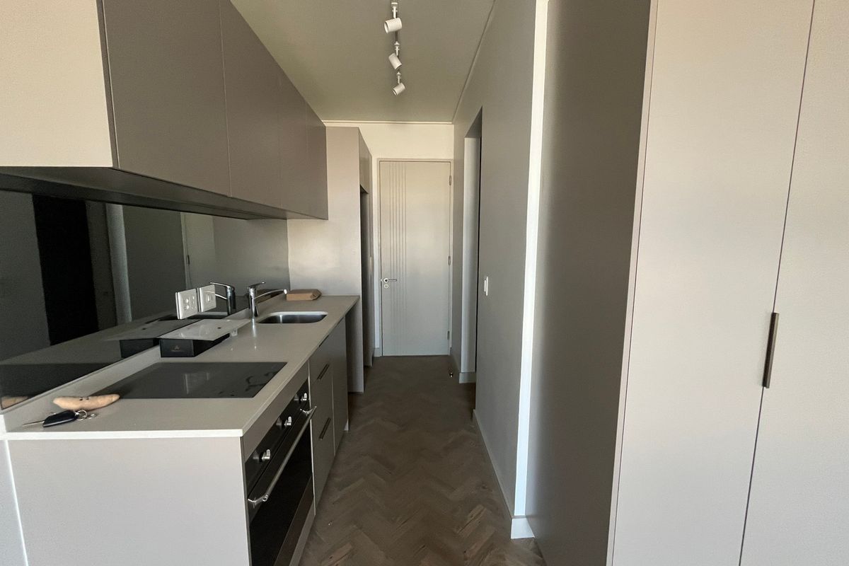Modern galley-style kitchen featuring sleek cabinetry, integrated appliances, mirrored splashback, and elegant herringbone timber flooring in a clean, minimalist space.
