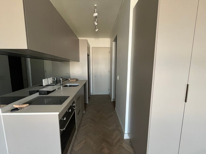 Modern galley-style kitchen featuring sleek cabinetry, integrated appliances, mirrored splashback, and elegant herringbone timber flooring in a clean, minimalist space.