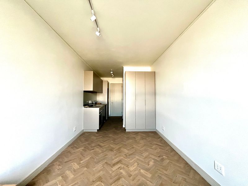 A bright, modern open-plan living space featuring high-quality herringbone timber flooring, track lighting, and sleek built-in cabinetry with integrated kitchen and storage.