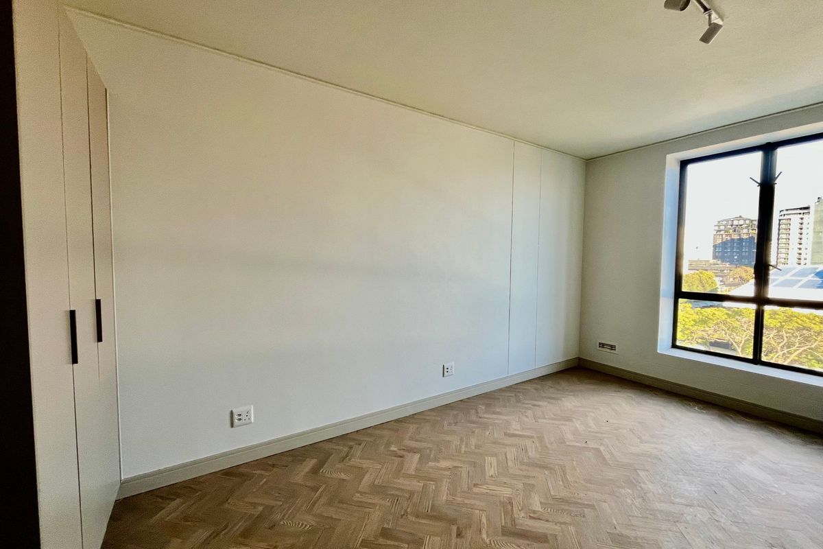 A bright, empty bedroom featuring elegant herringbone timber flooring, built-in wardrobes, and a large window offering natural light and urban views.
