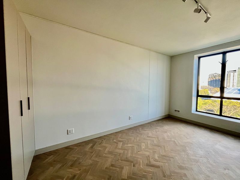 A bright, empty bedroom featuring elegant herringbone timber flooring, built-in wardrobes, and a large window offering natural light and urban views.