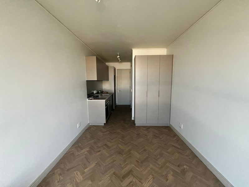 A modern open-plan studio apartment featuring high-quality herringbone timber flooring, sleek built-in kitchen cabinetry with integrated appliances, and a spacious built-in wardrobe.