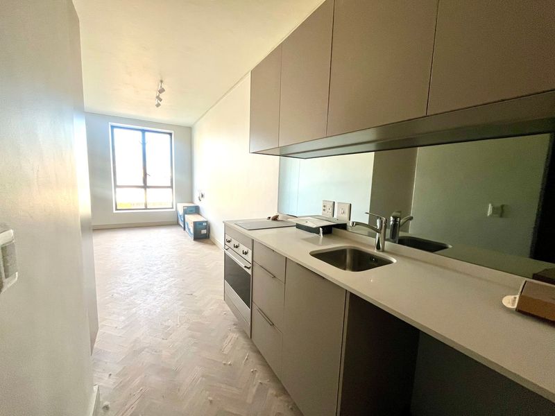 Modern open-plan kitchen and living area featuring sleek cabinetry, integrated appliances, mirrored splashback, and elegant herringbone timber flooring in a bright, vacant space.