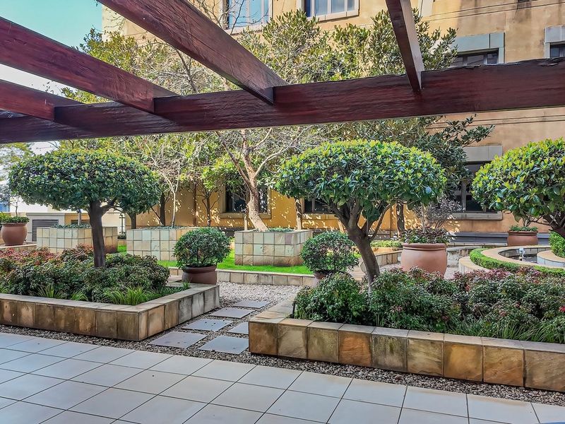 A well-maintained communal garden area featuring a wooden pergola, tiled patio space, and structured raised stone planters with established landscaping.
