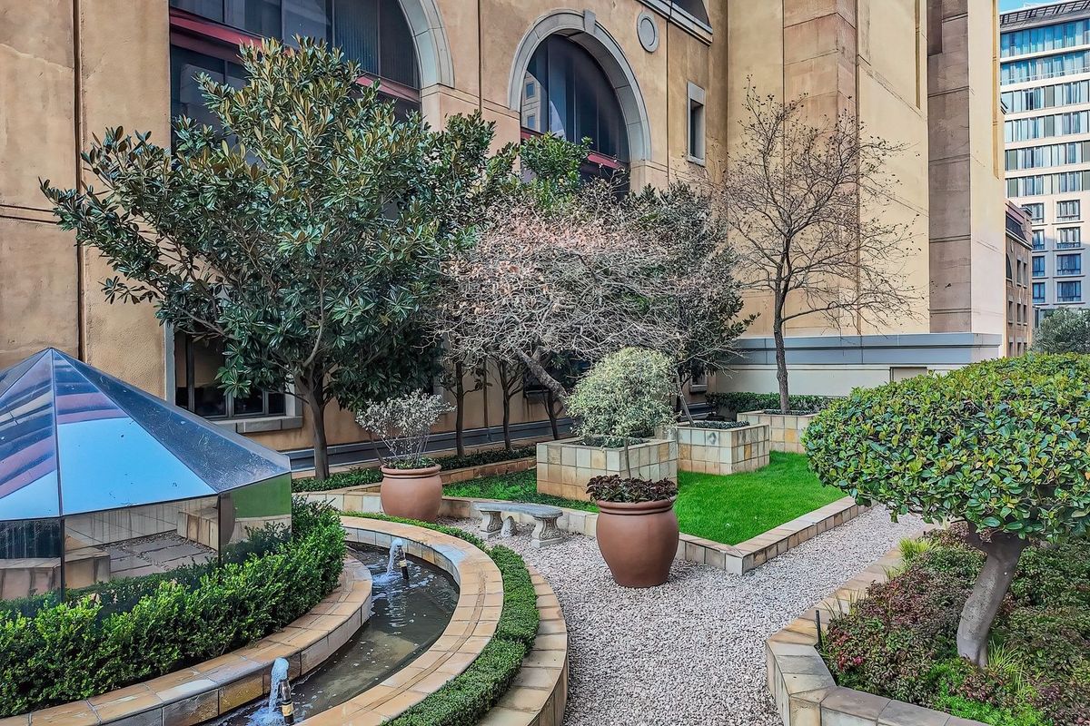 A well-maintained landscaped garden featuring gravel pathways, raised planter beds, a water feature, and a glass pyramid skylight set against the building facade.