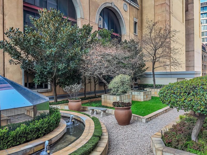 A well-maintained landscaped garden featuring gravel pathways, raised planter beds, a water feature, and a glass pyramid skylight set against the building facade.