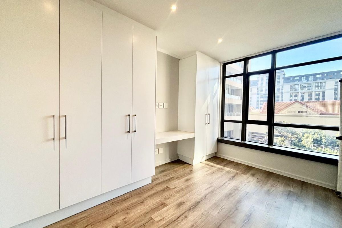 A bright, modern bedroom featuring extensive built-in cabinetry, a integrated desk, laminate flooring, and large windows offering natural light.