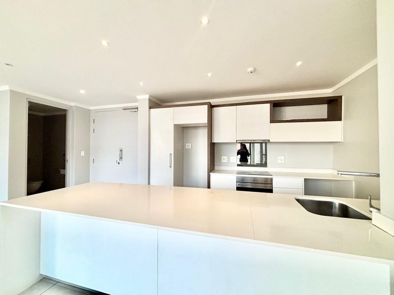 Modern open-plan kitchen featuring sleek white cabinetry, stone countertops, integrated oven, and recessed lighting in a clean, minimalist apartment setting.