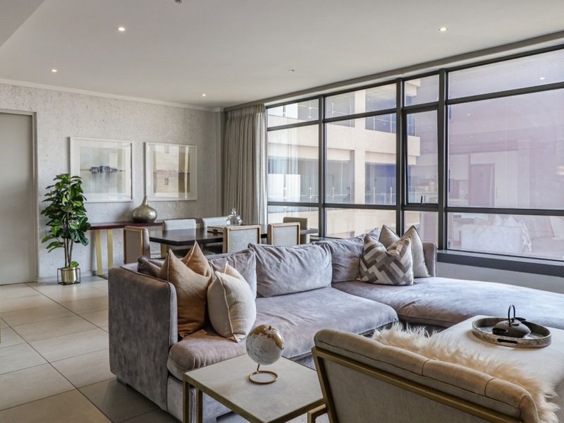 A bright open-plan living and dining area featuring large floor-to-ceiling windows, recessed downlights, and neutral tiled flooring.