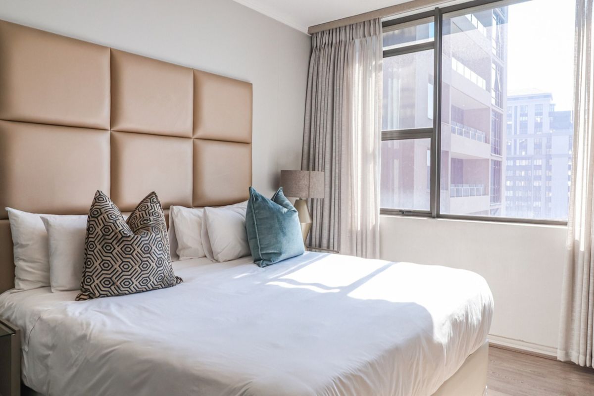 A well-maintained bedroom featuring a large window with curtains, an upholstered headboard, and laminate flooring, offering a bright and comfortable living space.