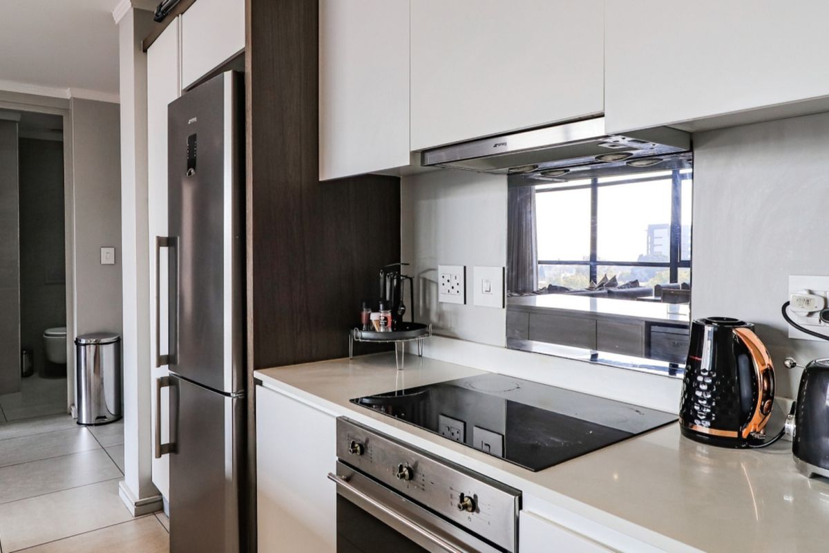 Modern kitchen featuring sleek white cabinetry, stone countertops, integrated oven and hob, and a stylish mirrored splashback, with a view into the adjacent hallway.