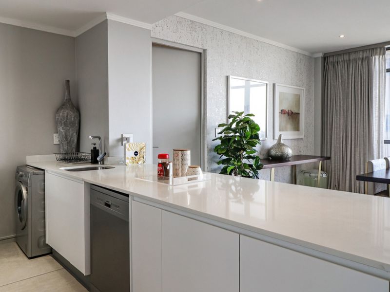 Modern open-plan kitchen area featuring sleek white cabinetry, stone countertops, integrated dishwasher, and tiled flooring, seamlessly flowing into the living space.