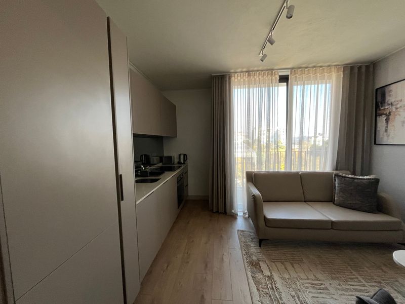 Modern open-plan living area featuring a sleek kitchenette with integrated appliances, wood-look flooring, and large sliding glass doors leading to a balcony.