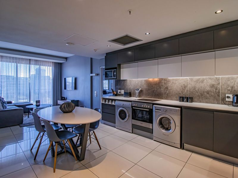 Modern open-plan apartment featuring a sleek kitchen with integrated appliances, stone backsplash, and high-gloss cabinetry, seamlessly flowing into the dining and living area.