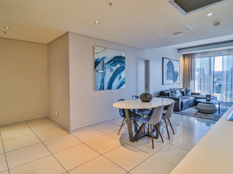 Modern open-plan living and dining area featuring polished porcelain tiles, recessed lighting, and floor-to-ceiling glass doors leading to a balcony.