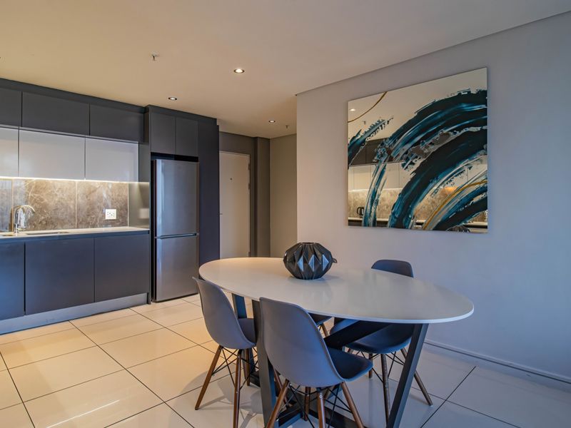 Modern open-plan kitchen and dining area featuring sleek dark cabinetry, stone splashback, tiled flooring, and recessed lighting in a contemporary apartment setting.