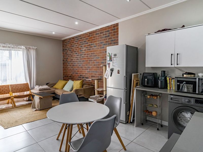 Open-plan living and kitchen area featuring a striking exposed brick wall, modern tiled flooring, and functional built-in cabinetry with integrated laundry plumbing.