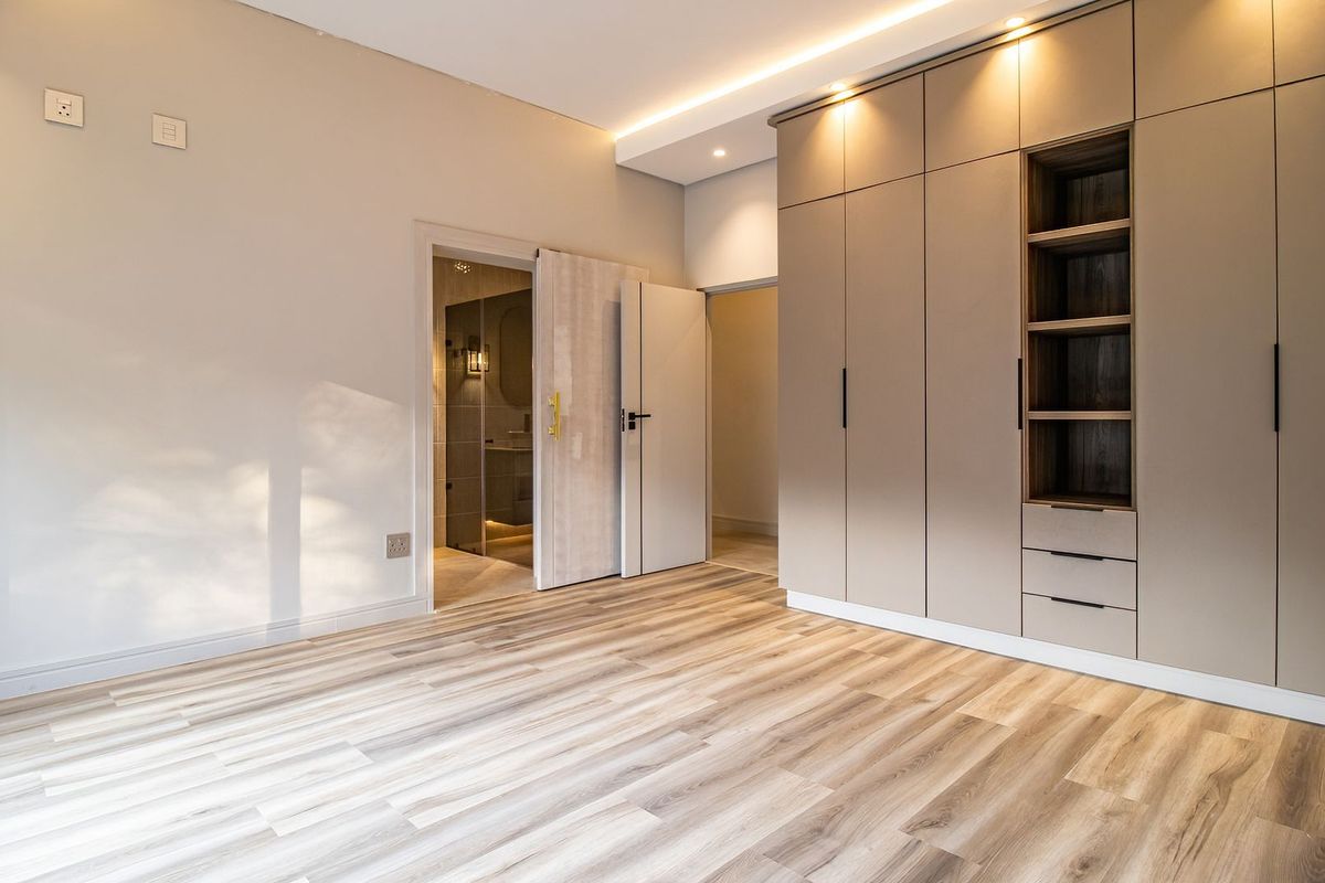 A modern, well-lit bedroom featuring high-quality laminate flooring, extensive built-in cabinetry with integrated shelving, and recessed ceiling lighting.