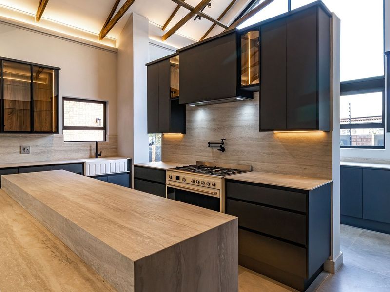 Modern kitchen featuring dark cabinetry, stone countertops, a farmhouse sink, gas stove with pot filler, and high ceilings with exposed timber rafters.