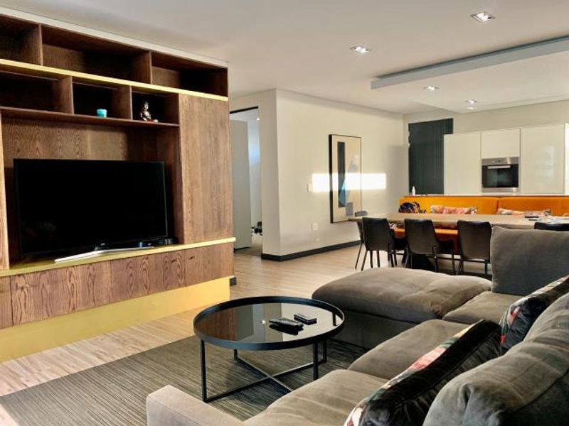 Modern open-plan living area featuring a large built-in wooden media unit, recessed lighting, and a seamless transition to the kitchen and dining space.