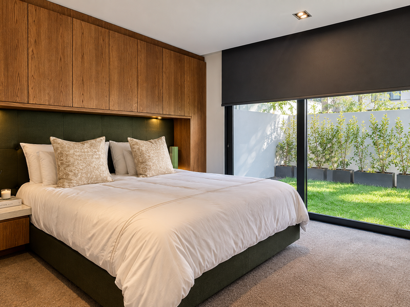 Modern bedroom featuring extensive built-in timber cabinetry, integrated headboard lighting, and large sliding glass doors opening to a private garden area.