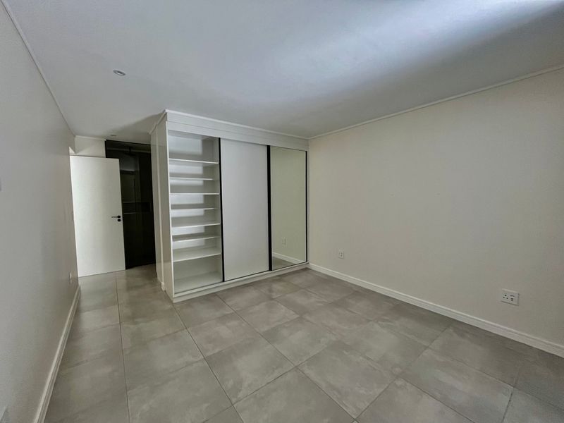 A clean, empty bedroom featuring large format floor tiles and a modern built-in wardrobe with mirrored sliding doors and open shelving.