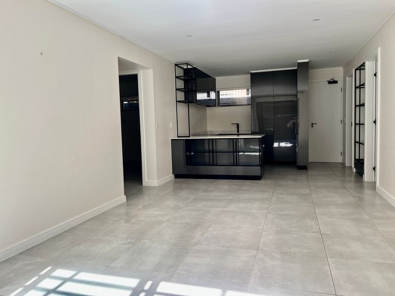 Modern open-plan living area featuring sleek dark cabinetry, a central kitchen island, and neutral tiled flooring throughout this contemporary apartment space.