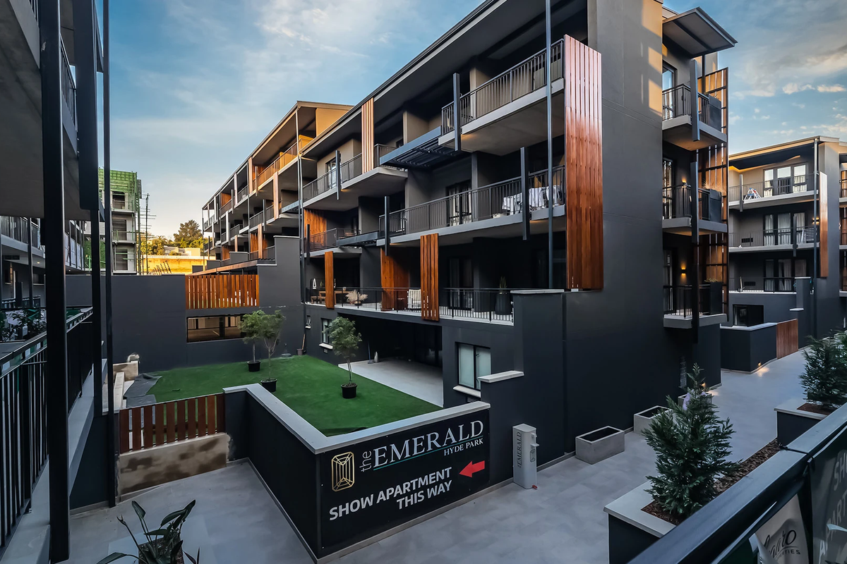 Modern apartment complex exterior featuring multi-level balconies, timber accents, and a landscaped courtyard area with paved walkways.