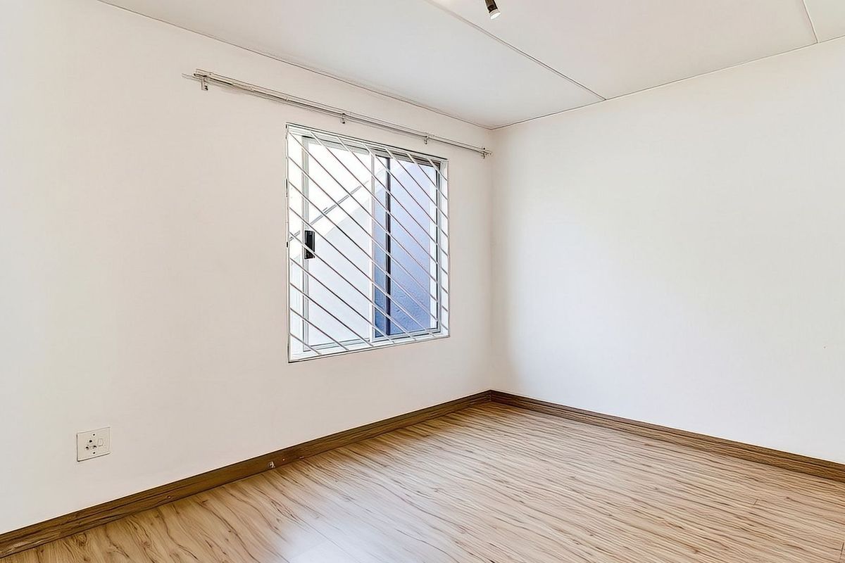A bright, empty bedroom featuring light wood-look laminate flooring, white walls, and a window fitted with security bars.