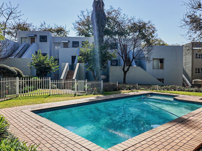 A well-maintained communal swimming pool area featuring brick paving, landscaped garden surrounds, and secure metal perimeter fencing within an apartment complex.
