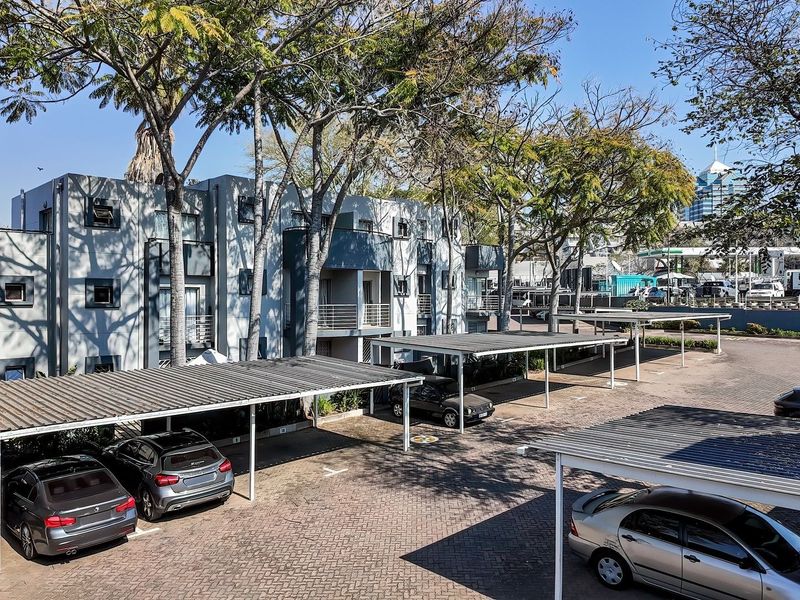 Exterior view of an apartment complex featuring covered parking bays, paved driveway, and multi-story residential units with balconies.