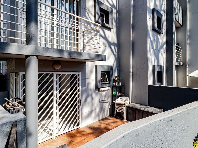Rear exterior view of an apartment building featuring a ground-level tiled patio, a first-floor balcony with security railing, and a water storage tank.