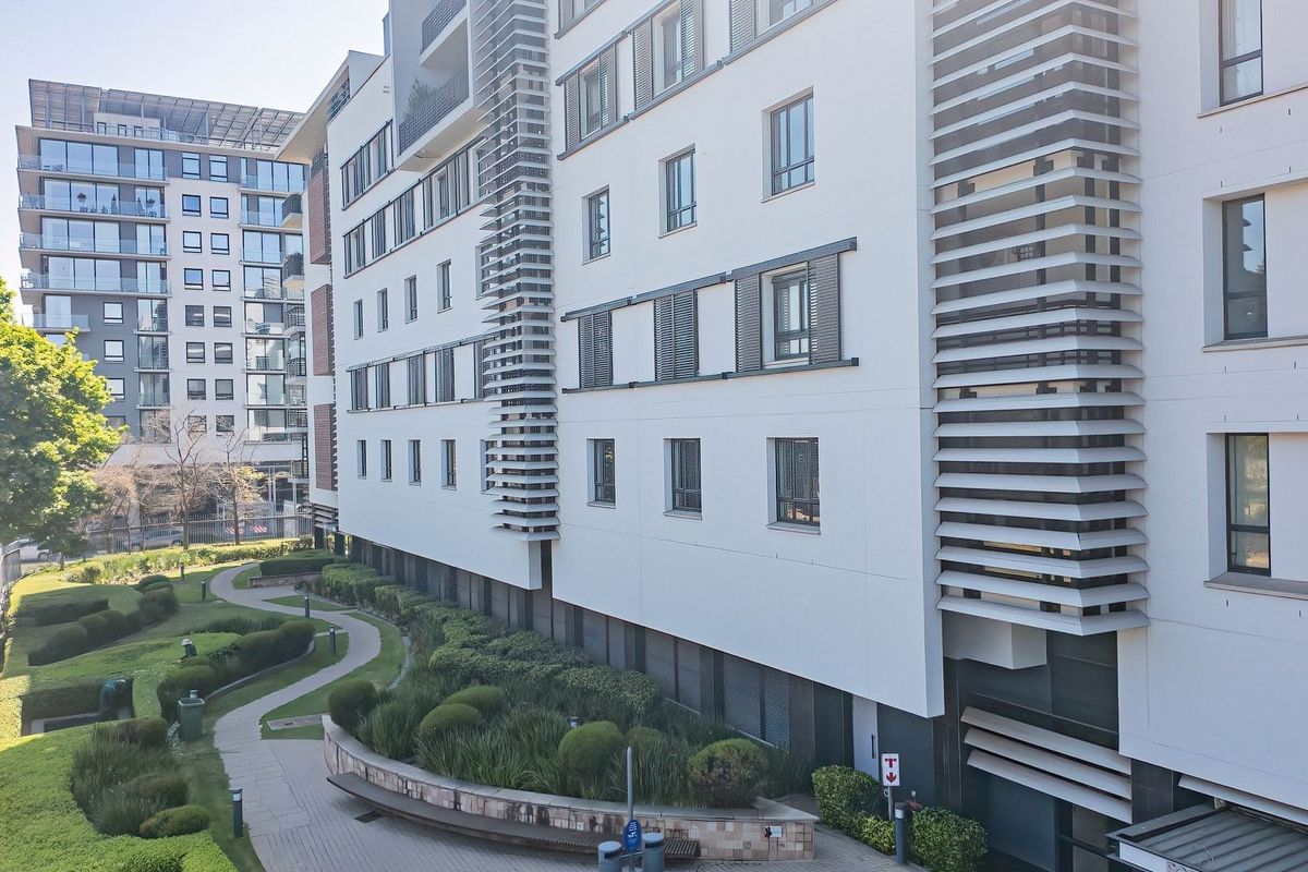An elevated view of the apartment building exterior featuring modern architectural facade detailing and a well-maintained landscaped garden with paved walkways.