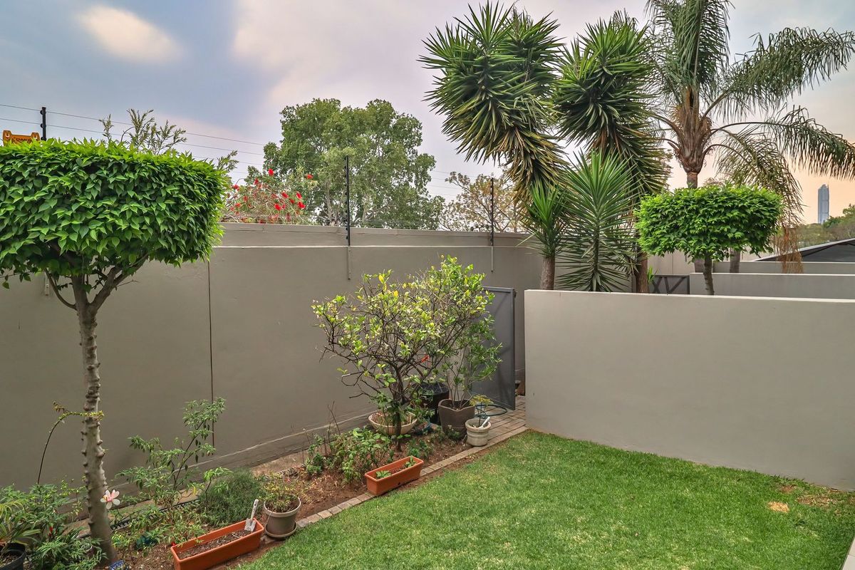 A well-maintained private rear garden featuring a manicured lawn, established trees, paved walkway, and secure boundary wall with electric fencing.