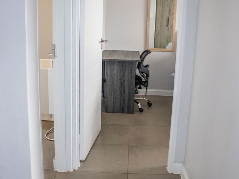 A view through a hallway leading into a bedroom or office space, featuring neutral tiled flooring and clean white door frames.
