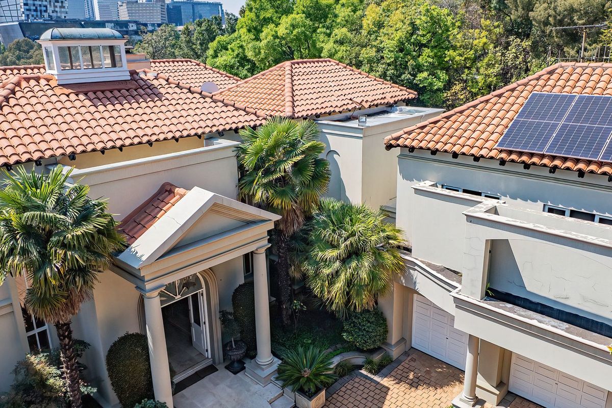 Aerial view of a cluster home featuring a terracotta tiled roof, solar panels, a classic portico entrance, and a paved driveway with double garage.