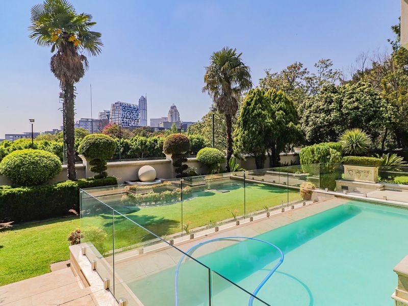 A beautifully landscaped garden featuring a swimming pool with a modern glass balustrade, stone water feature, and scenic city skyline views.